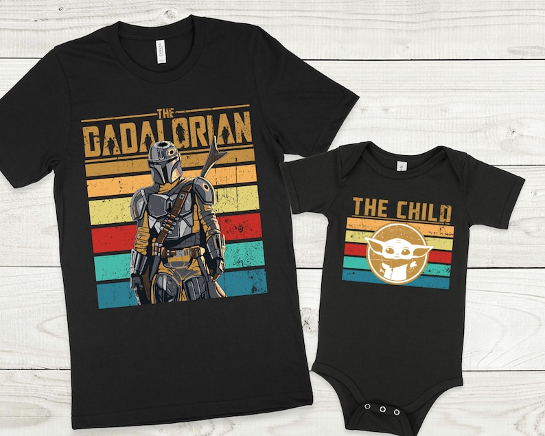 May include: Two black t-shirts with a retro design. The larger shirt features a Mandalorian character with the text "The Mandalorian" and a colorful striped background. The smaller shirt features a Baby Yoda character with the text "The Child" and a colorful striped background.