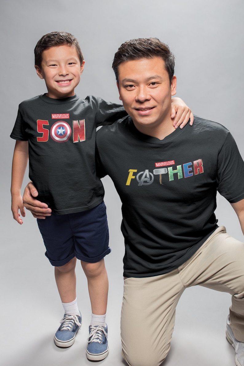 May include: A father and son wearing matching black t-shirts with the words "Marvelous Son" and "Marvelous Father" printed on them. The shirts feature various Marvel superhero logos and symbols.