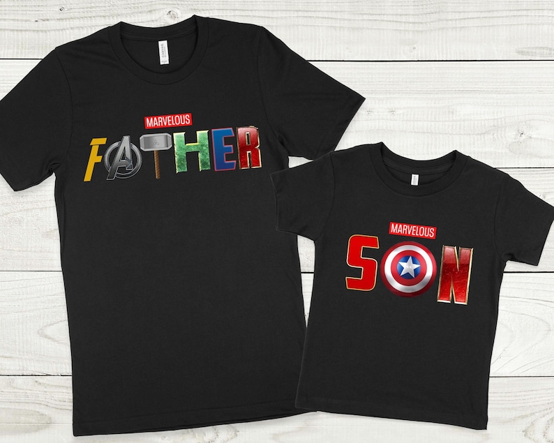 May include: Two black t-shirts with a Marvel theme. The larger shirt says "MARVELOUS FATHER" with the letters replaced by Marvel character symbols. The smaller shirt says "MARVELOUS SON" with the letters replaced by Marvel character symbols.