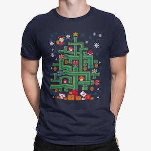 May include: Navy blue t-shirt featuring a pixelated Christmas tree design inspired by the Super Mario Bros. video game. The tree is composed of green pipes, red question mark boxes, and various characters and icons from the game, with snowflakes and a star.