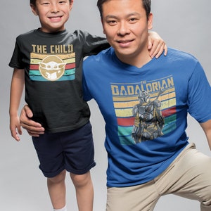 May include: A father and son wearing matching t-shirts. The son's shirt is black with the text "THE CHILD" and a retro-style image of a green alien. The father's shirt is blue with the text "THE  DADALORIAN" and a retro-style image of a Mandalorian.