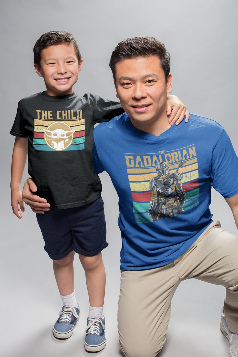 May include: A father and son wearing matching t-shirts. The son's shirt is black with the text "THE CHILD" and a retro-style image of a green alien. The father's shirt is blue with the text "THE  DADALORIAN" and a retro-style image of a Mandalorian.