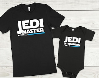 May include: Black t-shirt with white text that reads "Jedi Master" and a blue lightsaber graphic. A black baby onesie with white text that reads "Jedi in Training" and a blue lightsaber graphic.