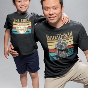 May include: A father and son wearing matching black t-shirts with a retro design. The father's shirt features the text "The Gadalorian" with a Mandalorian character. The son's shirt features the text "The Child" with a Baby Yoda character.
