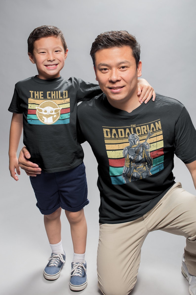 May include: A father and son wearing matching black t-shirts with a retro design. The father's shirt features the text "The Gadalorian" with a Mandalorian character. The son's shirt features the text "The Child" with a Baby Yoda character.