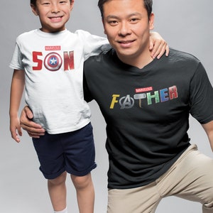 May include: A father and son wearing matching t-shirts with Marvel superhero logos. The son's shirt says "Marvelous SON" with a Captain America shield logo. The father's shirt says "Marvelous FATHER" with logos from Iron Man, Thor, and Captain America.