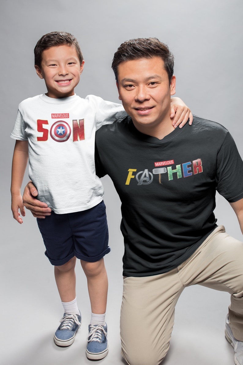 May include: A father and son wearing matching t-shirts with Marvel superhero logos. The son's shirt says "Marvelous SON" with a Captain America shield logo. The father's shirt says "Marvelous FATHER" with logos from Iron Man, Thor, and Captain America.