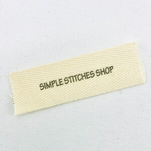 May include: A rectangular, off-white fabric label with the text "SIMPLE STITCHES SHOP" printed in dark green. The label has a textured weave pattern and is slightly angled against a white background.