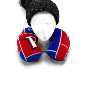 May include: A black knit beanie with a fluffy pom-pom sits atop a white mannequin head. A red, white, and blue travel neck pillow is positioned below the head. The pillow has a team logo design.