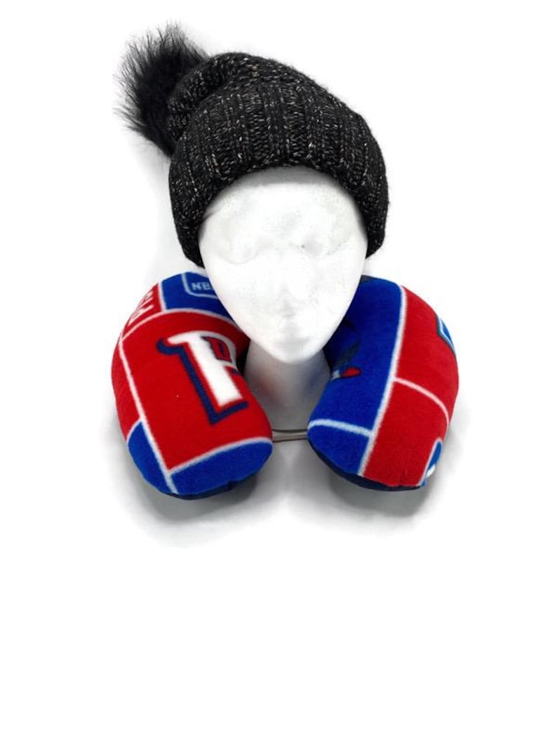 May include: A black knit beanie with a fluffy pom-pom sits atop a white mannequin head. A red, white, and blue travel neck pillow is positioned below the head. The pillow has a team logo design.
