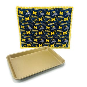 May include: A gold-colored rectangular baking sheet with rounded corners. A blue and yellow patterned kitchen towel featuring the text "Michigan" and the letter "M" is in the background. The towel has a yellow gingham border.