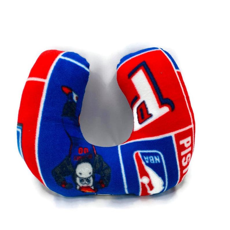 May include: A blue and red U-shaped travel pillow featuring the Detroit Pistons logo and NBA branding. The pillow has a blue background with a graphic of a basketball player. The pillow is designed for neck support.