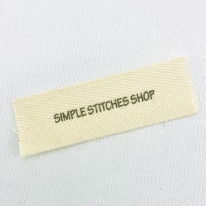 May include: A rectangular, off-white fabric label with the text "SIMPLE STITCHES SHOP" printed in dark green. The label has a textured weave and is slightly angled against a white background.