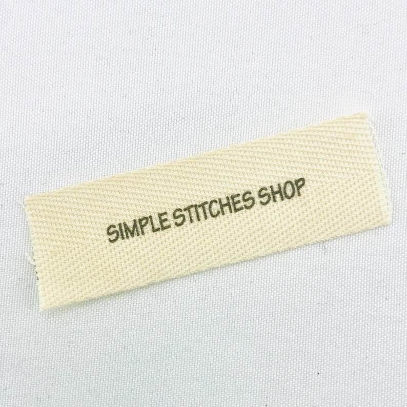 May include: A rectangular, off-white fabric label with the text "SIMPLE STITCHES SHOP" printed in dark green. The label has a textured weave and is slightly angled against a white background.