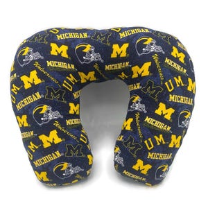 May include: A navy blue travel neck pillow with a repeating pattern of yellow "M" logos, football helmets, and the word "Michigan". The pillow is designed for comfort and support during travel.