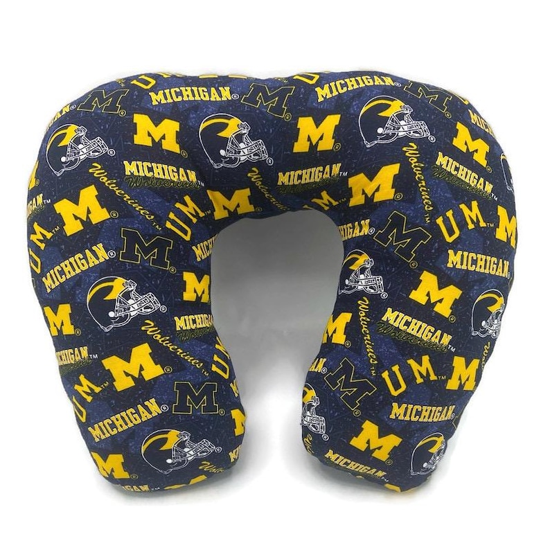 May include: A navy blue travel neck pillow with a repeating pattern of yellow "M" logos, football helmets, and the word "Michigan". The pillow is designed for comfort and support during travel.