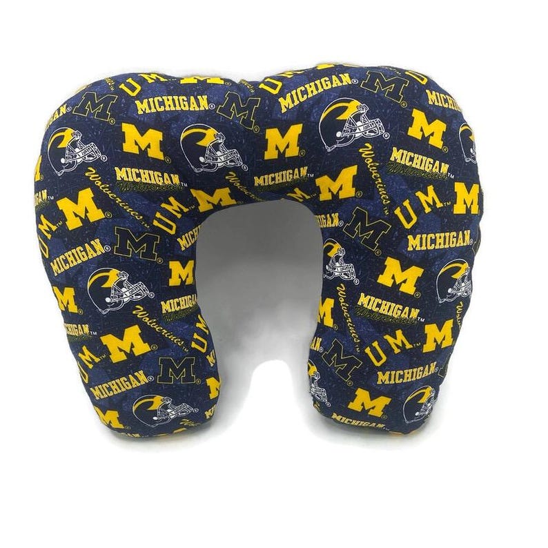 May include: A navy blue travel pillow with a repeating pattern of yellow "M" logos, football helmets, and the word "MICHIGAN". The pillow is designed to support the neck and head, ideal for travel or relaxation.