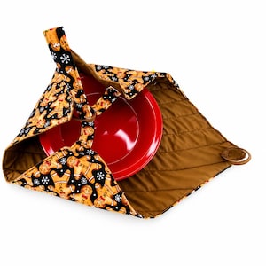 May include: A quilted fabric bowl cozy featuring a black background with a repeating pattern of gingerbread men and white snowflakes, containing a bright red bowl. The interior of the cozy is a solid brown fabric with diagonal stitching.