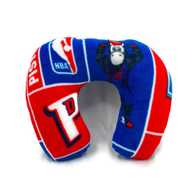 May include: A blue, red, and white travel pillow featuring the Detroit Pistons logo and a cartoon gorilla wearing a jersey. The pillow is shaped like a horseshoe and has the NBA logo.