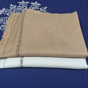 May include: Two rectangular fabric wraps, one in tan and one in white, featuring a woven texture and a decorative border with a small geometric pattern near the fringed edges. The wraps are folded and stacked on top of each other.