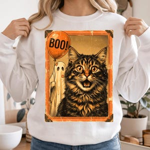 May include: A white long-sleeve sweatshirt featuring a rectangular graphic print of a brown tabby cat with wide eyes and an open mouth, standing next to a white ghost holding an orange balloon with the text BOO! printed in black letters.