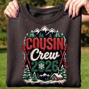 May include: A dark charcoal gray sweatshirt featuring a graphic with the text "COUSIN Crew 2026". The design includes red knitted-style lettering, white script, green numbers, a white snowflake, snowy mountains, pine trees, a cabin, and red ski poles.