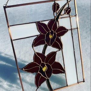 May include: A rectangular stained glass suncatcher featuring two dark red orchid flowers with yellow centres and small buds on a stem. The design is set against a background of clear and opaque white glass panels, all framed in a thin copper metal border.