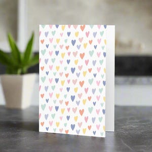 May include: A folded greeting card with a white background covered in a pattern of small, colorful hearts. The hearts are in shades of pink, blue, yellow, and green. The card is standing upright on a dark surface.