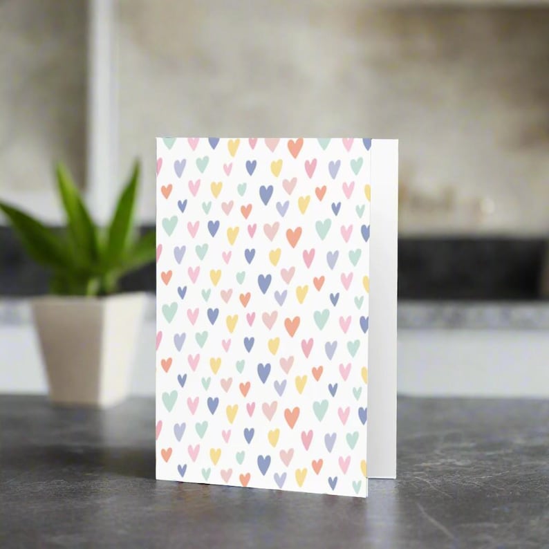 May include: A folded greeting card with a white background covered in a pattern of small, colorful hearts. The hearts are in shades of pink, blue, yellow, and green. The card is standing upright on a dark surface.