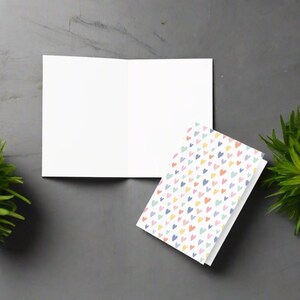 May include: An open, blank white greeting card next to a stack of cards with a colorful heart pattern. The hearts are in shades of pink, yellow, blue, and green. The cards are on a gray surface.