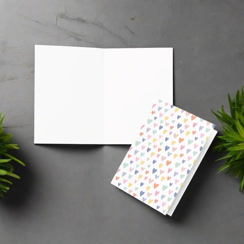 May include: An open, blank white greeting card next to a stack of cards with a colorful heart pattern. The hearts are in shades of pink, yellow, blue, and green. The cards are on a gray surface.