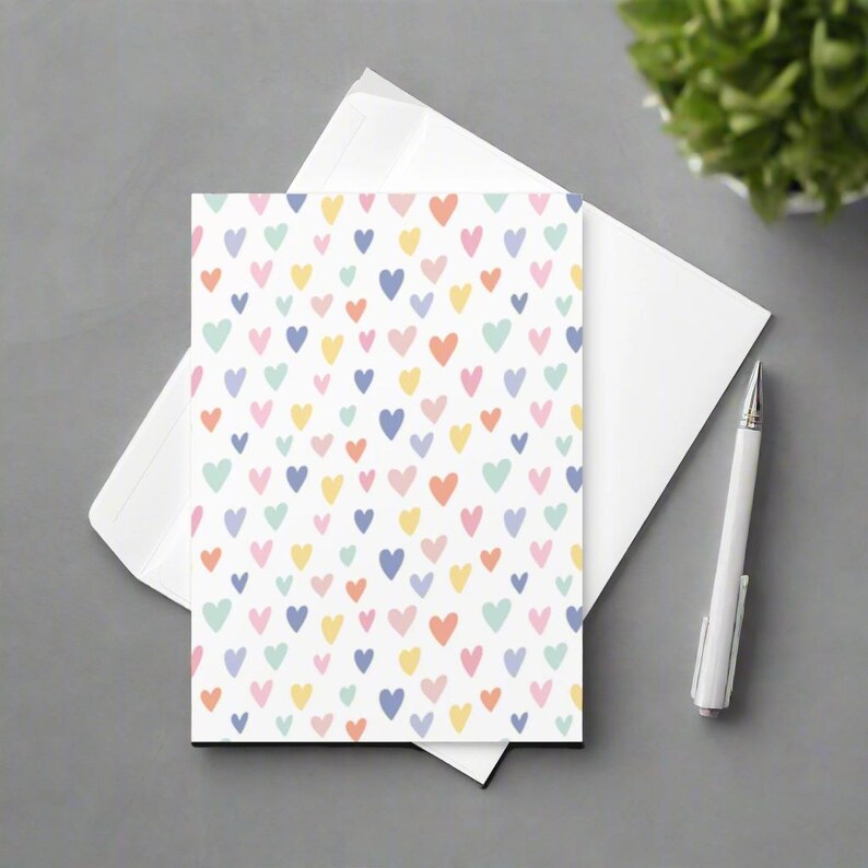 May include: A white greeting card with a pattern of colorful hearts in pastel shades of pink, yellow, blue, and orange. The card is placed on top of a white envelope, with a white pen resting nearby. The card and envelope are on a gray surface.