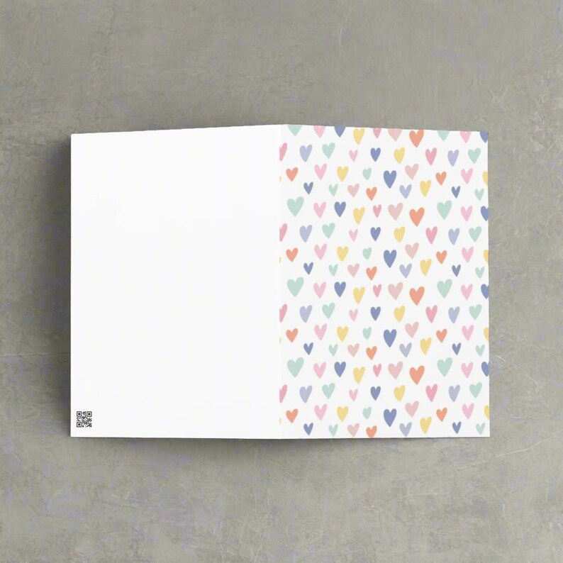 May include: A folded greeting card with a white interior and a colorful heart pattern on the right side. The hearts are in various pastel colors, including pink, yellow, blue, and green. A QR code is in the lower left corner.