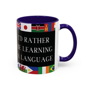 May include: White ceramic mug with a navy blue interior and handle. The mug features the text "I'D RATHER BE LEARNING A LANGUAGE" in white, set against a black background. Flags of various countries are displayed around the rim.