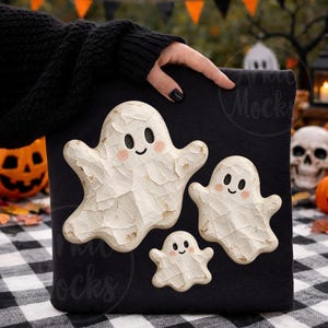 May include: A black sweatshirt featuring a graphic print of three white ghosts with textured, carved-look surfaces, black oval eyes, and small pink cheeks. The ghosts are arranged in a cluster, with one large, one medium, and one small ghost.