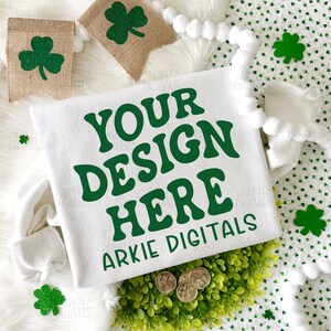 May include: White sweatshirt with the text "YOUR DESIGN HERE" in green, along with "ARKIE DIGITALS". The image includes shamrock decorations, burlap banners with shamrocks, and gold coins. The design is perfect for St. Patrick's Day.