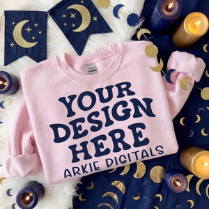 Puede incluir: Una sudadera rosa claro con el texto azul marino "YOUR DESIGN HERE". La sudadera está rodeada de decoraciones celestiales azul marino y doradas, incluyendo pancartas y adornos en forma de luna. Hay velas alrededor de la sudadera.