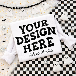 May include: White sweatshirt with the text "YOUR DESIGN HERE" in black, with "Arkie Mocks" below. The sweatshirt is surrounded by Halloween decorations, including ghost-shaped ornaments and a black and white checkered pattern.
