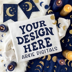 Puede incluir: Una sudadera blanca con el texto "YOUR DESIGN HERE" en azul marino. La imagen incluye decoraciones con temática de luna azul marino y dorada, velas y una pancarta. El texto "ARKIE DIGITALS" también es visible.
