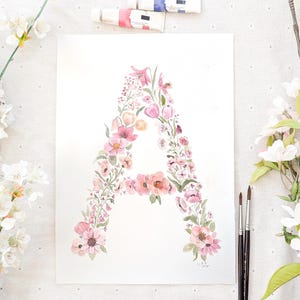 May include: Watercolor painting of the letter "A" formed by pink and peach flowers and green leaves. The artwork is surrounded by white flowers, paint tubes, and paint brushes. The floral design is on a white background.
