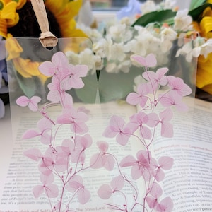 May include: Clear, rectangular bookmark with pressed pink hydrangea flowers. A beige ribbon is attached for use. The floral design adds a touch of nature to reading materials.