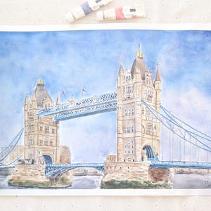 May include: A watercolor painting of London's Tower Bridge featuring two stone towers connected by a blue suspension span against a light blue sky. The artwork displays detailed architectural elements, including arched entryways and pointed roofs.