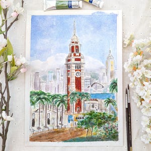 Könnte beinhalten: Aquarellmalerei einer Stadtlandschaft mit einem hohen rot-weißen Uhrturm, Palmen und anderen Gebäuden. Das Kunstwerk ist vor einem blauen Himmel mit weißen Wolken platziert. Farbtuben und Pinsel sind sichtbar.