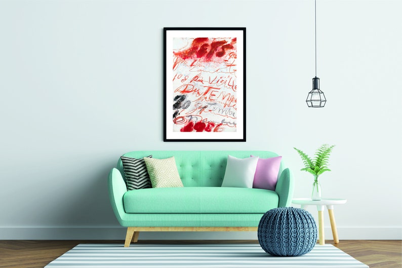 May include: A framed abstract art print with red and black brushstrokes and handwritten text in French. The print is hanging on a light blue wall above a teal sofa with pillows and a blue knitted pouf.