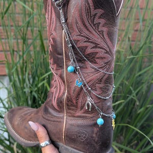 Op de afbeelding: Een bruine leren westernlaars met rode decoratieve stiksels en een zilverkleurige metalen kettingaccessoire aan de zijkant. De ketting bevat kleine turquoise kralen, een blauwe vlinderbedel, een hoefijzer en een gele bliksemschicht.