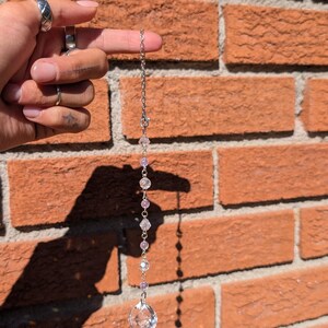 Peut inclure: Un attrape-soleil suspendu composé d'une chaîne en métal argenté alternant des perles de cristal facettées transparentes et de petites perles sphériques violet clair, se terminant par un grand pendentif en cristal transparent à facettes.