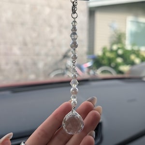 Peut inclure: Un ornement de voiture suspendu composé d'une chaîne argentée avec une alternance de perles de cristal facettées transparentes et de perles blanches sphériques, se terminant par un grand pendentif en cristal sphérique à facettes.
