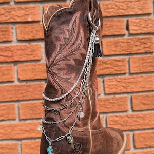 May include: A brown leather western boot featuring intricate red stitching and a decorative silver-toned chain accessory. The accessory includes multiple layered chains, a small black tassel, a turquoise lightning bolt charm, and a revolver charm.