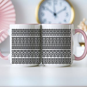 Pode incluir: Duas canecas de cer&acirc;mica brancas com al&ccedil;as e bordas rosa. Cada caneca apresenta um padr&atilde;o geom&eacute;trico preto de linhas e formas horizontais. Um rel&oacute;gio com moldura dourada &eacute; vis&iacute;vel ao fundo.