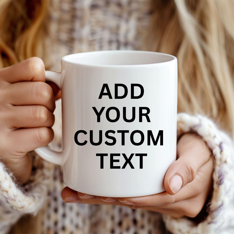 Pode incluir: Uma caneca de cer&acirc;mica branca com a frase "ADD YOUR CUSTOM TEXT" em preto, segurada por uma pessoa. A caneca tem uma al&ccedil;a e formato cil&iacute;ndrico. O fundo est&aacute; desfocado, mostrando uma pessoa com um su&eacute;ter.